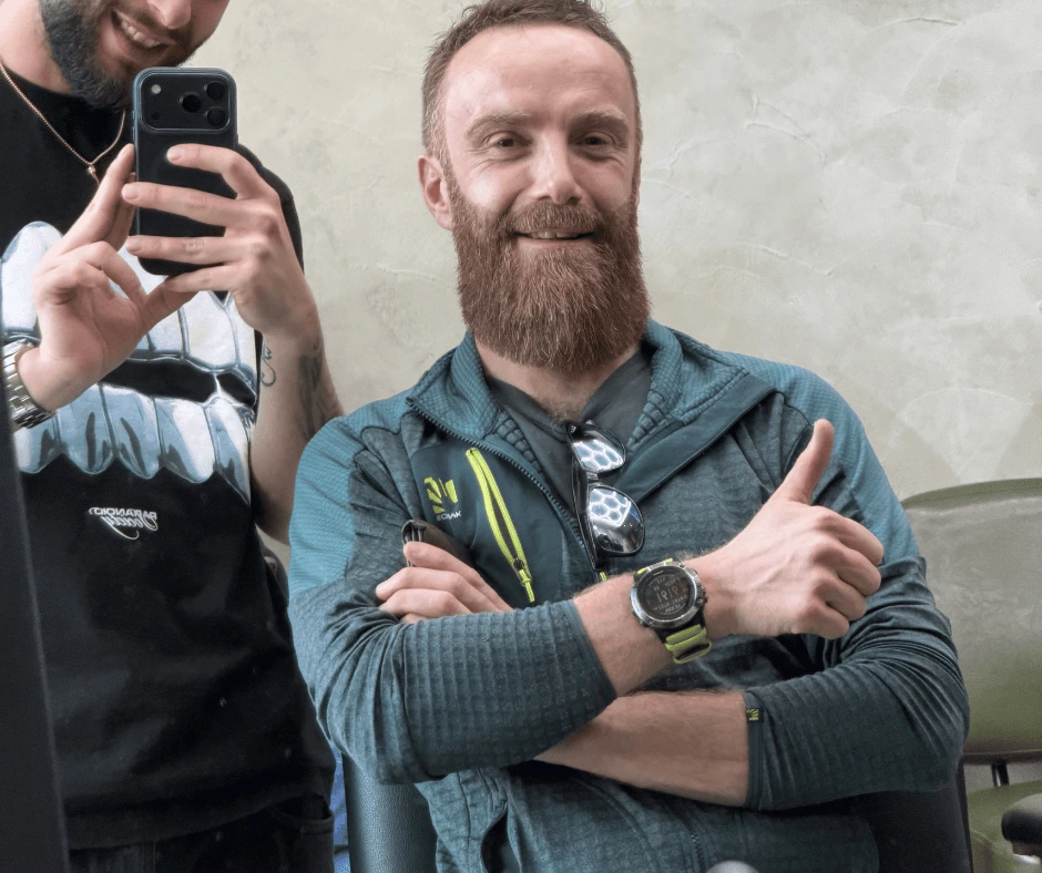 Cliente sorridente con barba rossa rifinita mostra il pollice in su accanto al barbiere